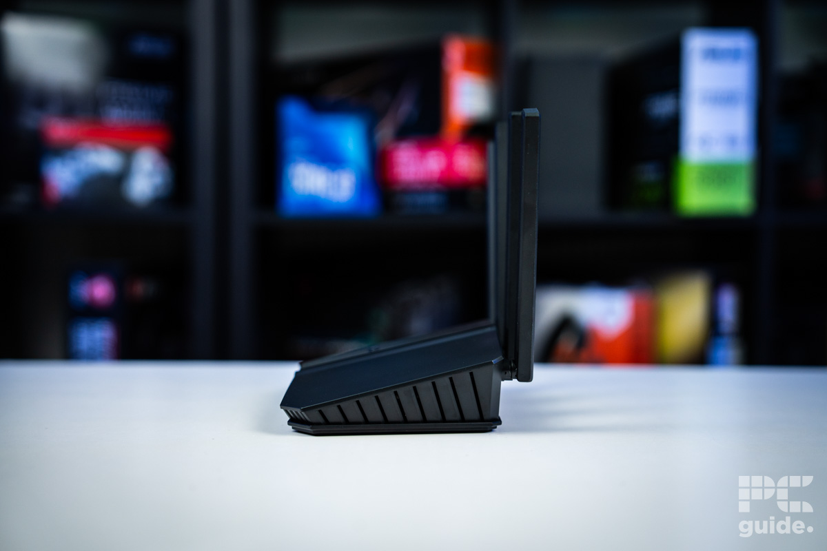 The black ASUS RT-BE58U (BE3600 router sitting on a white table with a background of blurry PC component boxes on a bookshelf. Its shot from the left side showing the slope of it and its side vents with the antenna behind it.