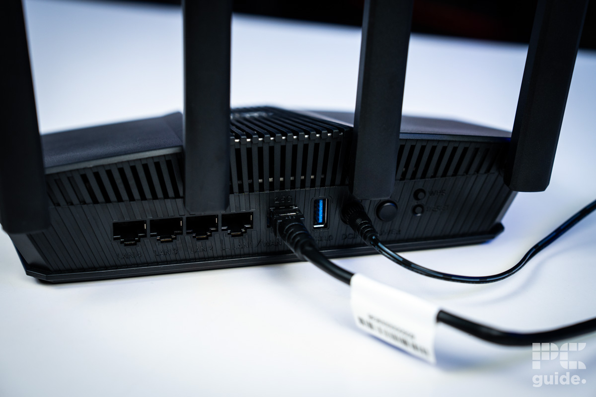 The back of the black ASUS RT-BE58U (BE3600 router sitting on a white table showing its back ports with the power and WAN port plugge din.