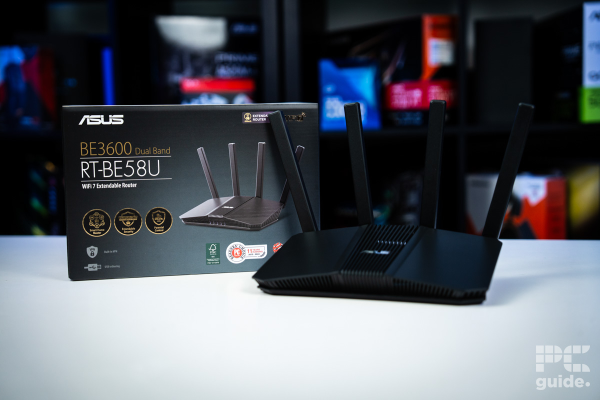 The black ASUS RT-BE58U (BE3600 router sitting on a white table with a background of blurry PC component boxes on a bookshelf. It sits in front of its box with four antenna sticking up from it.