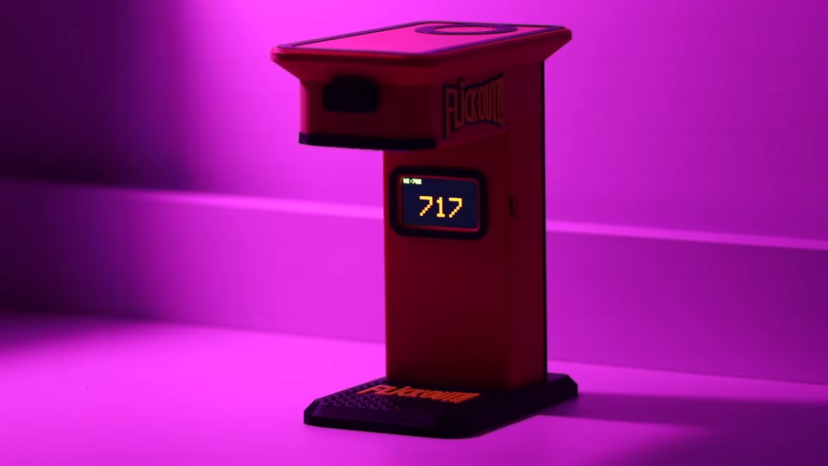 Flick-Out desktop game with a black base and red pillar and overhang. The screen reads 717 as it stands on a white table and with white background and is lit with a purple light.