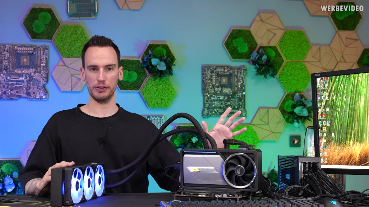 A screenshot from a der8auer video. It showcases him on the left side looking down the camera with an AIO cooler on the table in front of him, with a RTX 5090 AStral GPU on his right middle. On the furthest right a part of a monitor can be seen.