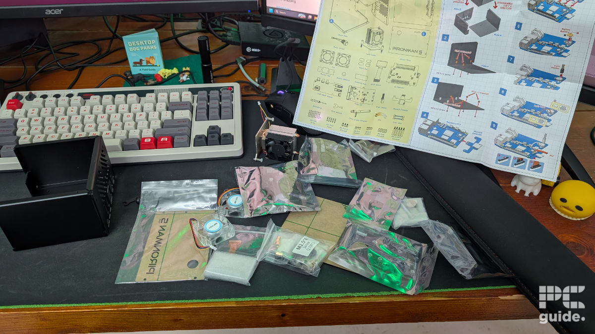 A desk with a mousepad and keyboard. In the main frame is the contents of the Pironman 5 case box. It consists of the two plate frame, a set of paper instructions, and several plastic baggies full of components, and two fans, and tower cooler.
