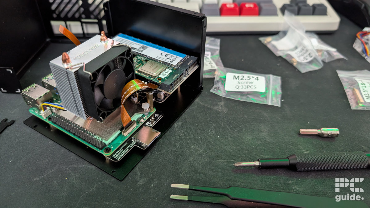 The Raspberry Pi 5 with a tower cooler installed on it. Along with a NVMe PIP next to it connected via a FCP cable. It's all seated on a plate of the Pironman 5 MAX case. On the right are some plastic baggies, screwdriver, and tweezers.