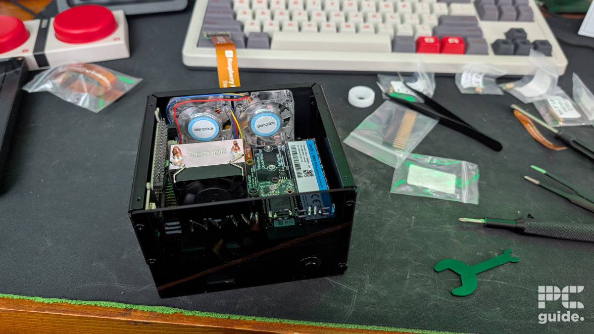 A mousepad with a keyboard in the background. in the forgeround is the Pironman 5 Max case with components built inside of it. prominently are the tower cooler, and fans behind, along with the NVMe and AI chip on the right. There are also baggies scattered on the right.