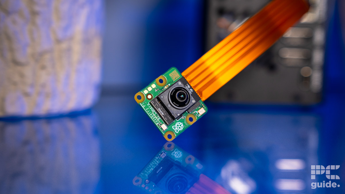 A bare PCB with a camera lens in the centre. Out of it is a golden ribbon cable towards the out of focus PC. It sits on a blue table and a jagged plant pot is on the left.