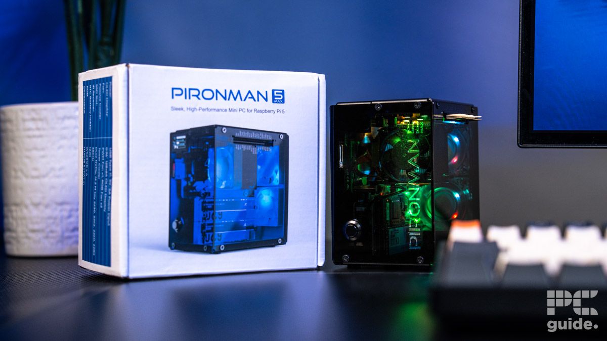The Pironman 5 Max box that's whtie and shows the case on the front. On the right is the cases itself built and glowing in lights. There is an out of focus keyboard on monitor peaking in from the right. In the lft background is a vase and plant.