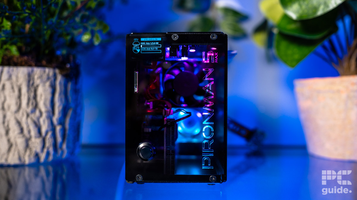 Pironman 5 Max case front with the name cutout down the side. A fan is seen trhough the middle panel. There is a push button in the bottom left. A screen readout can be seen in the top left reading CPU usage and temperature, RAM usage and disk space. In the background are two out of foucs plant pots on either side.