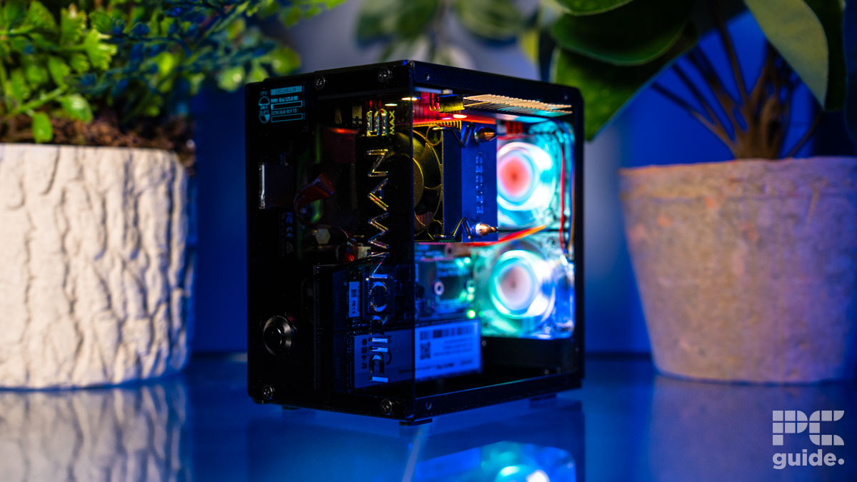 The Pironman 5 Max angled front. The insdie can be seen with lit up fans and coolers. Behind its got two plants as it stands on a blue table.