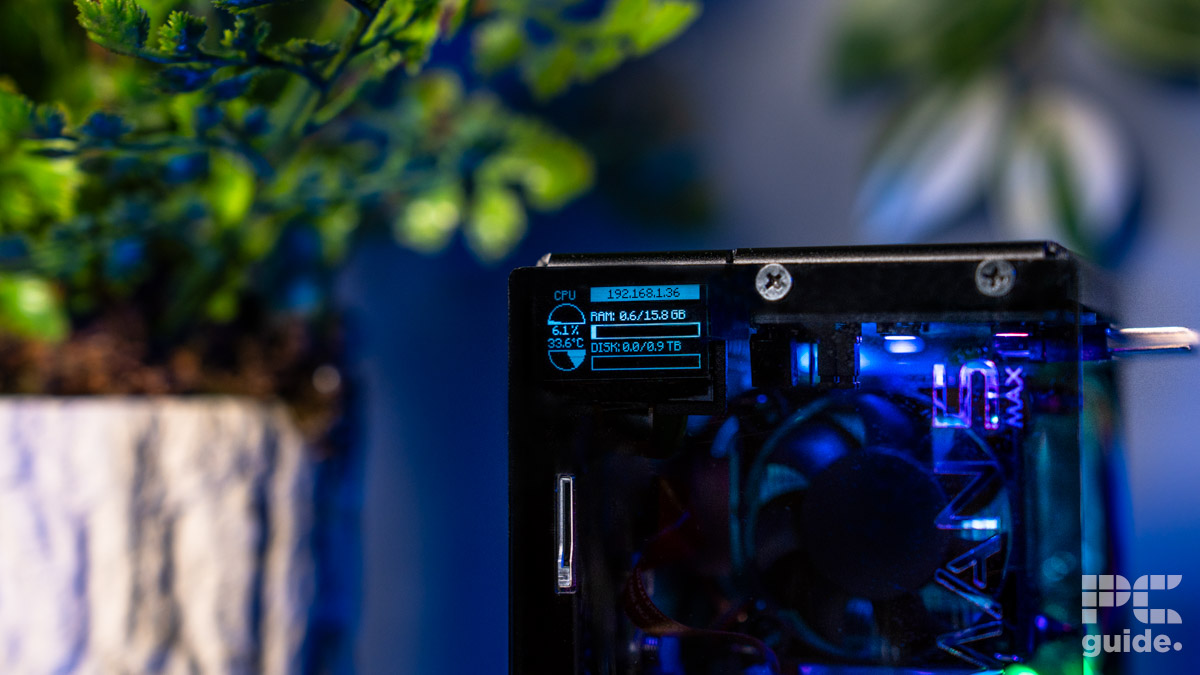 The top edge of the front of the Pironman 5 Max case. It shows two screws and the OLED screen in the top left just behind the panel. It reads CPU utilization, temperature, RAM and disk usage. On the left is an out of focus plant.