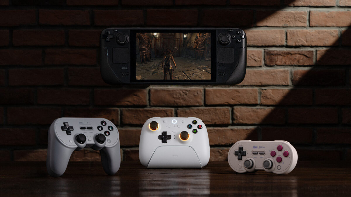 A brick wall background and wooden table at the bottom. On the brick wall is a floating Steam Deck with a game running on it. On the table are three 8Bitdo controllers