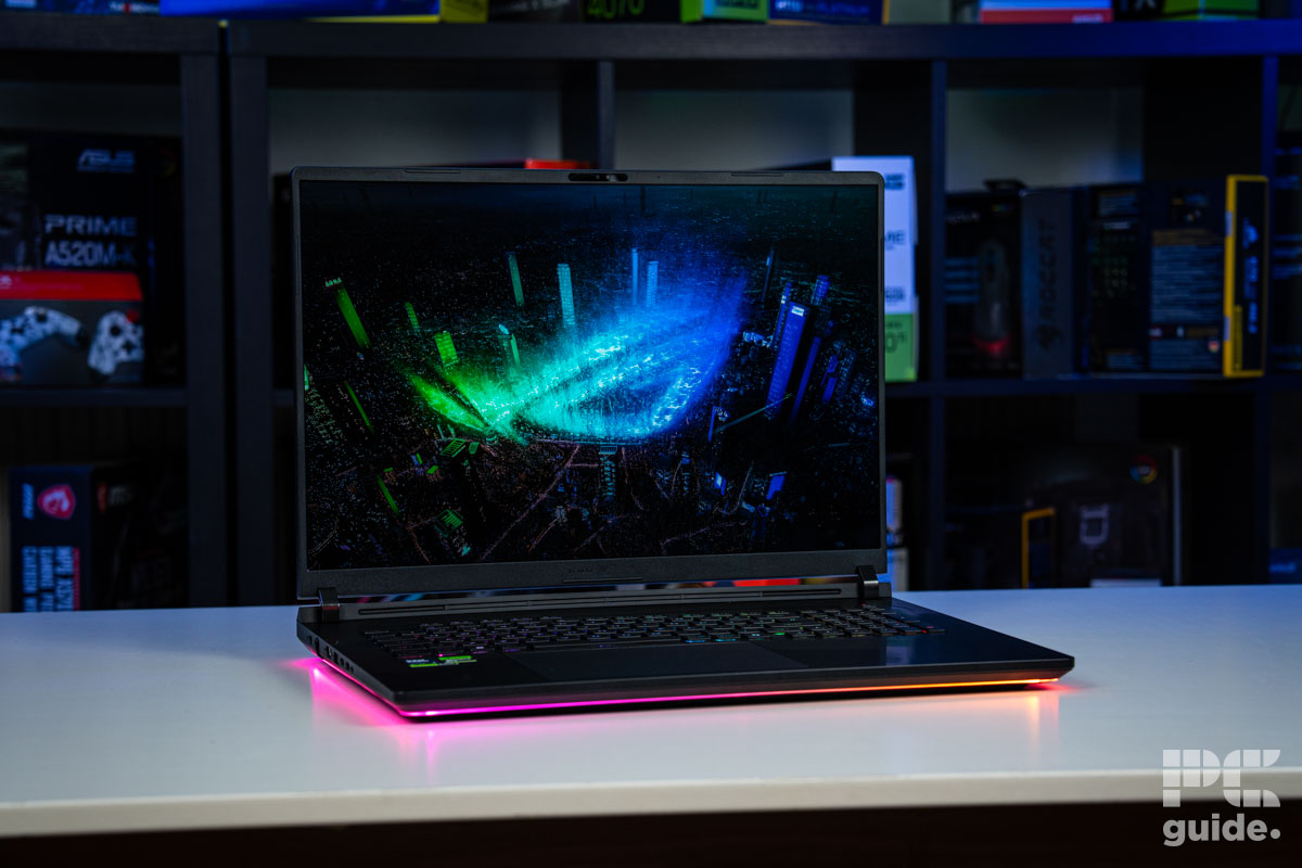 A photo of the ROG Strix Scar 18 G835LX, sitting on a white table, with a blurred background of PC component boxes on a bookshelf. It shows the laptop turned on and open. The light around the base colored purple, and the keyboard is lit up. The creen shows a cityscape with an ROG logo made of RGB shining in the middle.