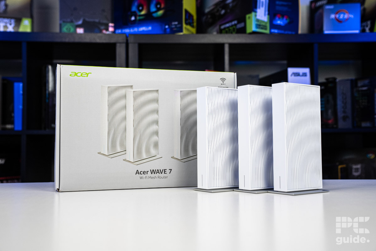 The three Acer Wave 7 routers standing next the their box sitting on a white table with a background of blurred PC component boxes on a bookshelf.