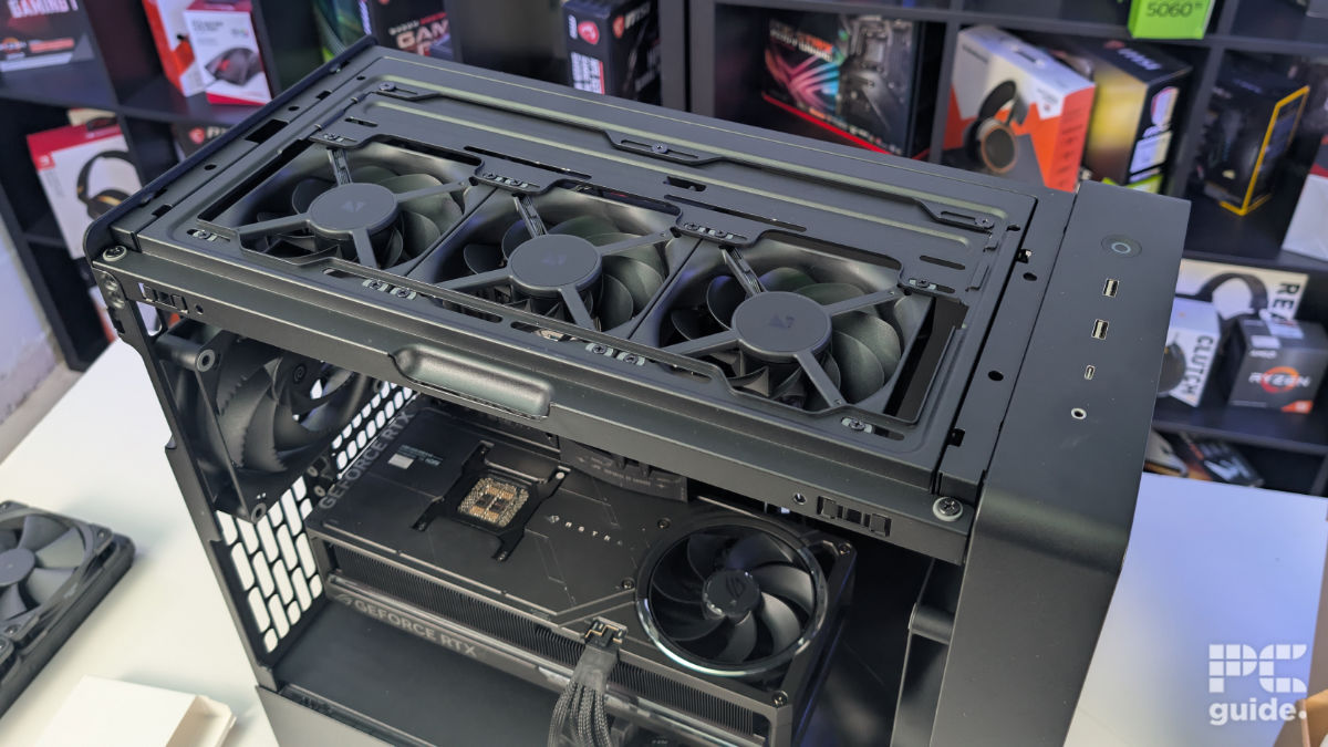 A top down view of the HAVN BF 360 case with the top taken off and three 120mm fans installed. With the inside built wihth PC components, standing on a white desk with a background of PC component boxes on a bookshelf.