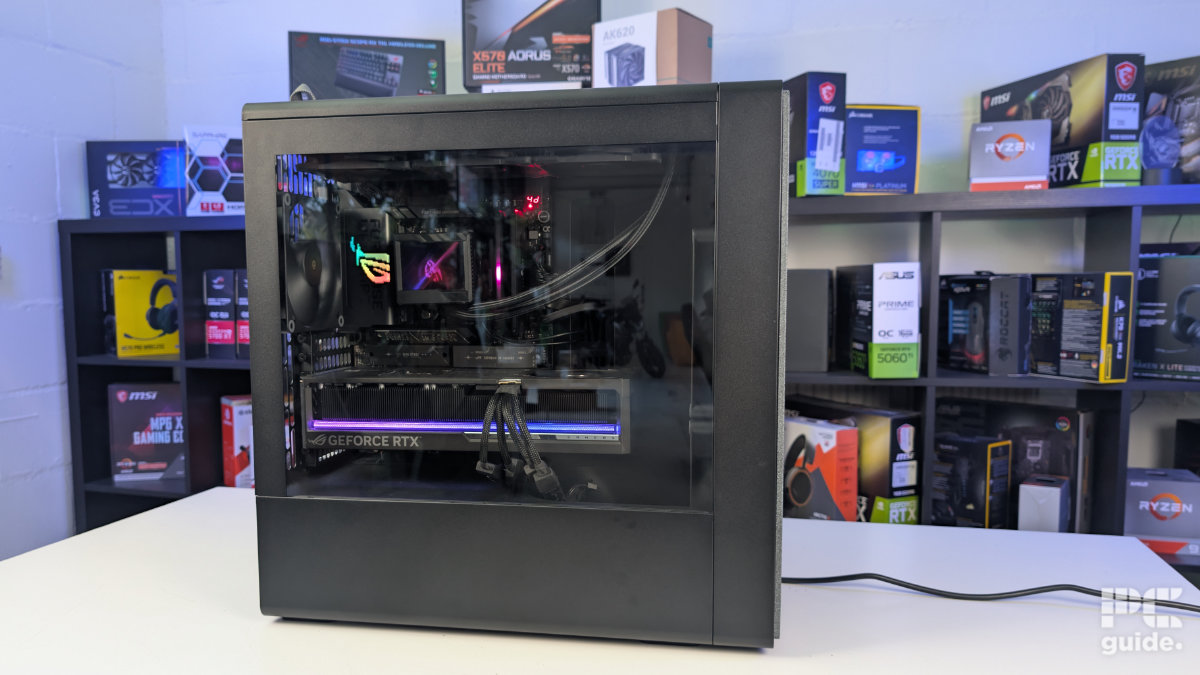 A view inside of the HAVN BF 360 standing on the white table and background of PC component boxes on a bookshelf. Inside the case is built wiht a GPU and AIO. The glass panel is closed and glares as the PC is on with lights shining inside.