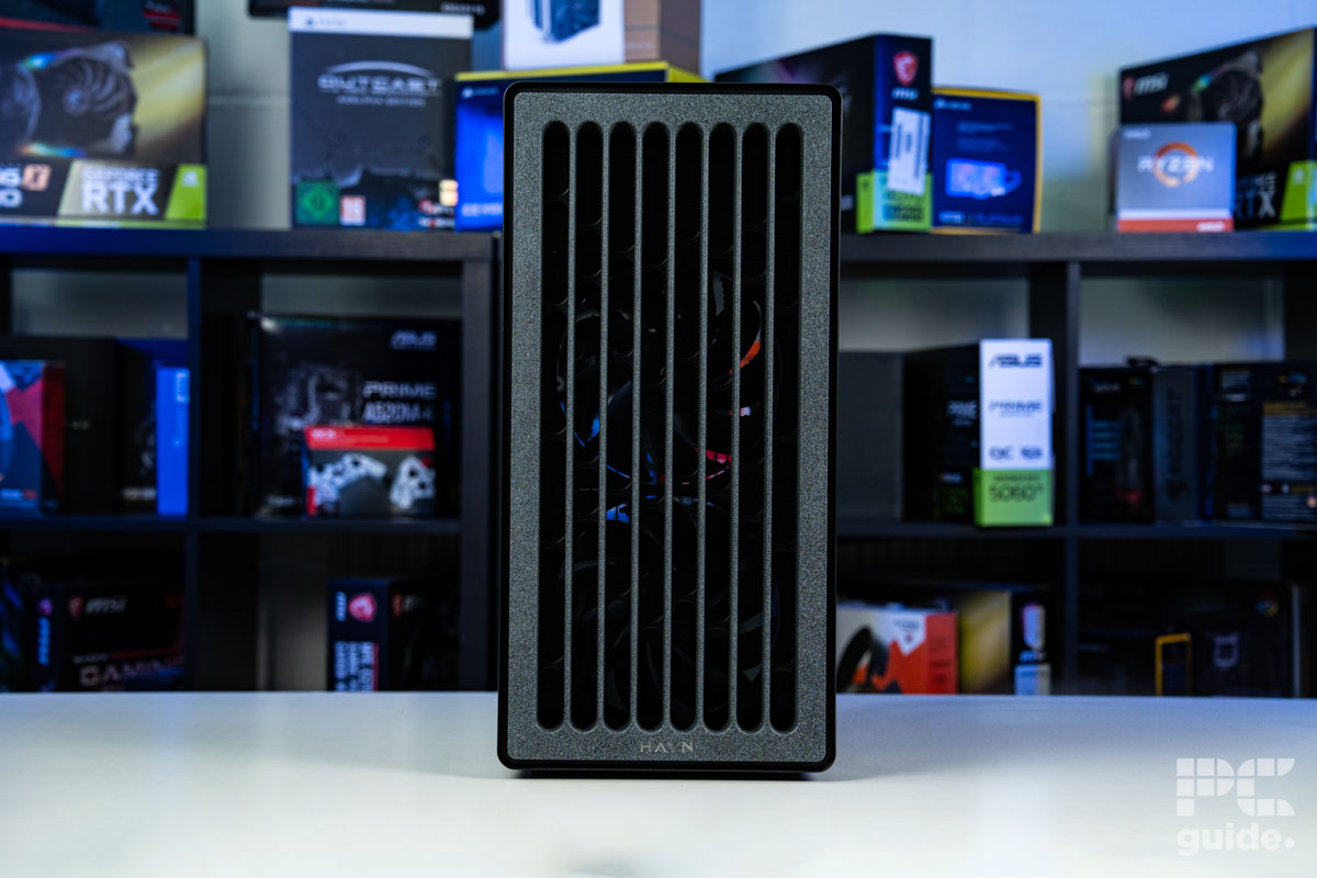 A shot of the fron of the HAVN BF 360 Flow case on a white table with a background of PC component boxes on a bookshelf. It has a faux stone looking design with vertical slits for airflow.