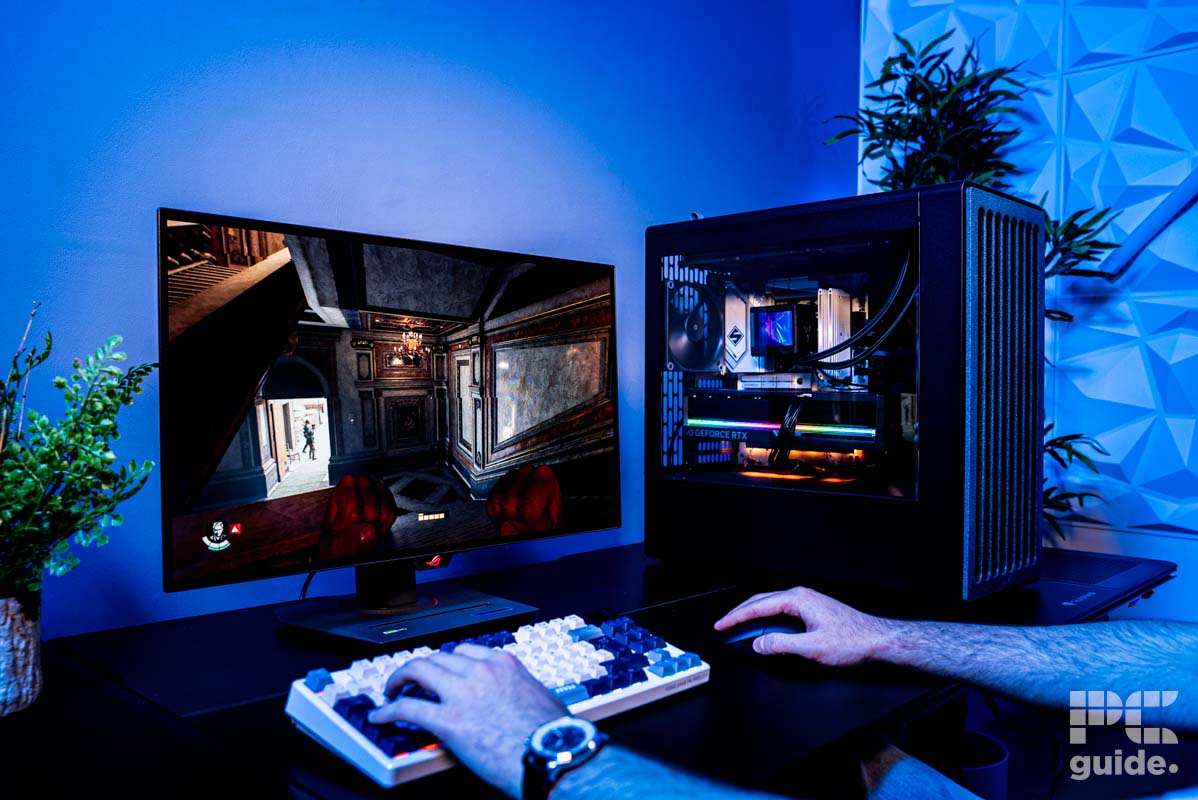The HAVN BF 360 Flow case on a table with a build inside. Next to it is a keyboard and mouse with hands using them to play Indiana Jones on the monitor. The background is a blue wall, with two plants on either side.