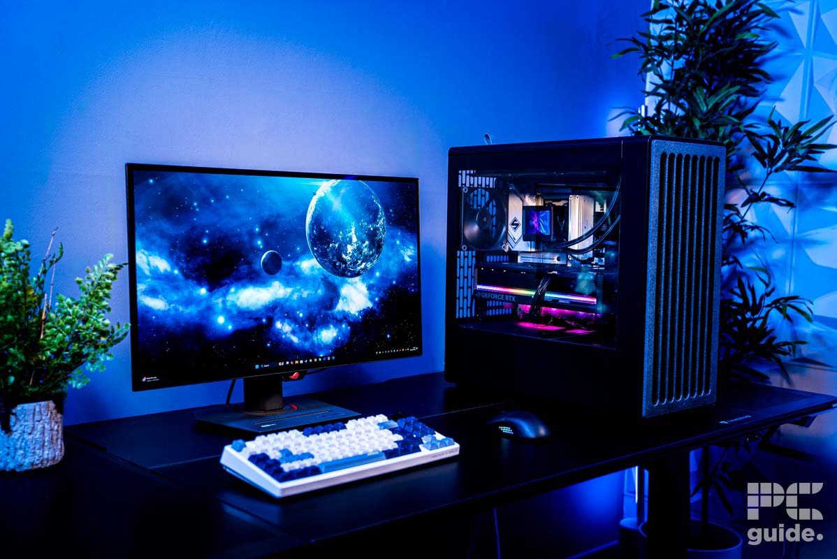 The HAVN BF 360 Flow case with a PC built inside full of color. It sits on a table, with a monitor, keyboard, and mouse next to it. Along with a two plants and a blue wall behind it.