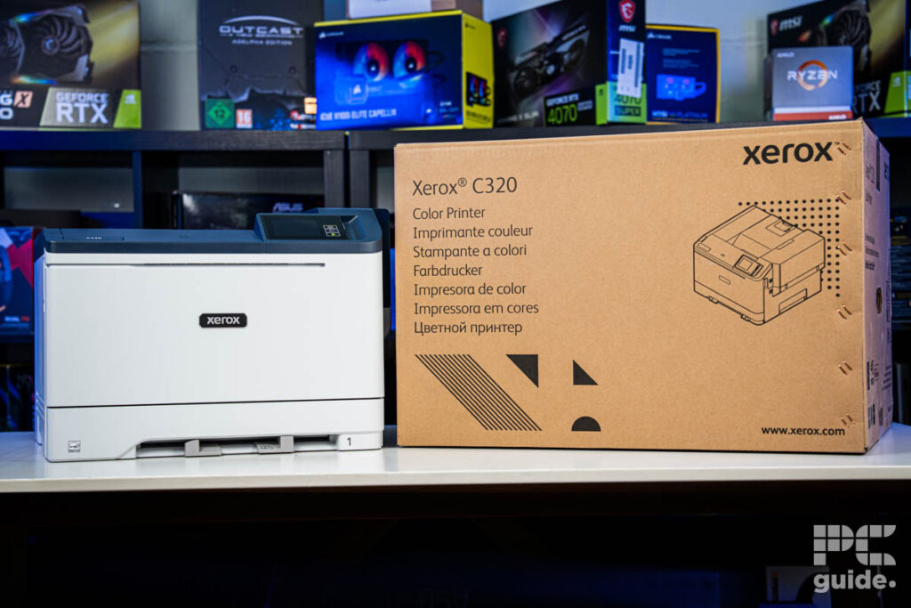 The Xerox C320 front on, on the left side, and its box on the right of it. Sitting on a white table, with a background of a bookshelf filled with PC component boxes.